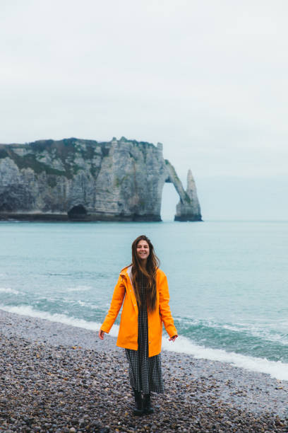 glückliche frau im regenmantel zu fuß am schönen strand mit natürlichen bogen in etretat, frankreich - women rear view one person arch stock-fotos und bilder