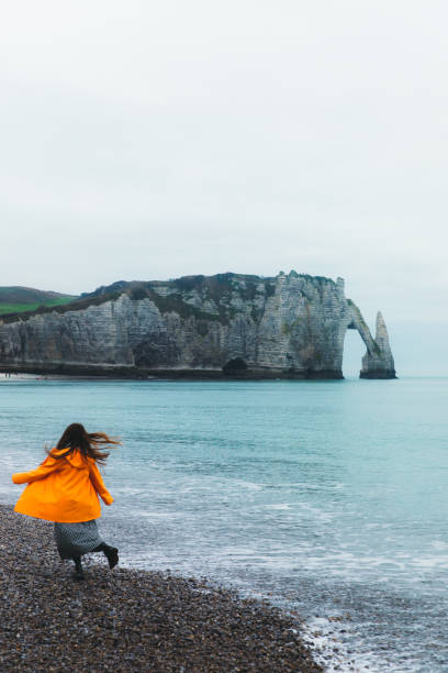 donna con l'impermeabile che cammina sulla bellissima spiaggia con arco naturale a etretat, francia - women rear view one person arch foto e immagini stock