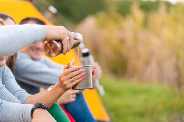 A group of friends is enjoying a warming drink from a thermos, on a cool evening by a fire in the forest. Fun time camping with friends.