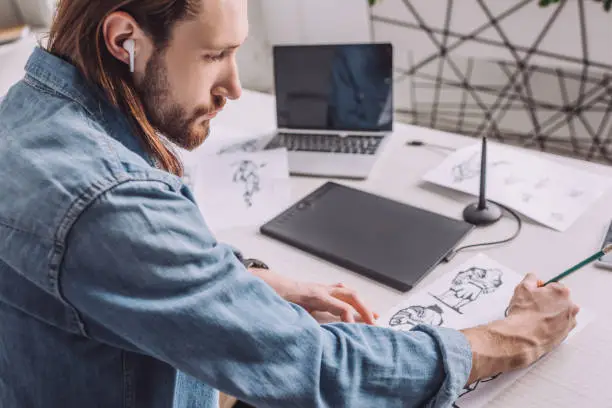 Photo of bearded of illustrator in wireless earphone drawing sketches