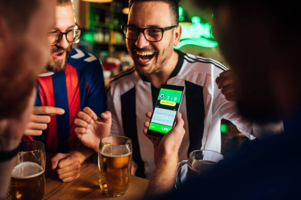 Friends drinking beer, watching soccer game and using mobile app for betting Soccer fans drinking beer at the pub and using mobile app for betting. friends in bar with phones stock pictures, royalty-free photos & images