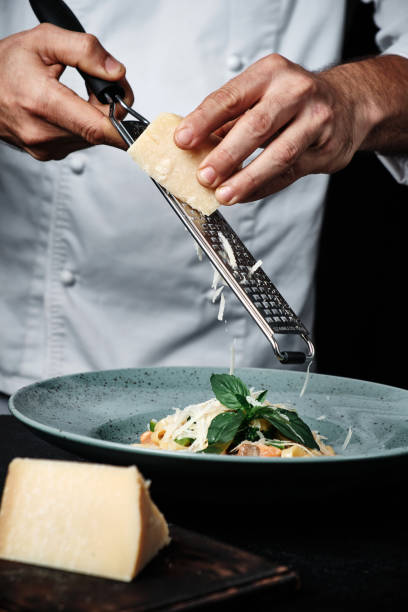 chef mani cucinando pasta italiana e aggiungendo parmigiano di formaggio in piatto su sfondo nero - grater foto e immagini stock
