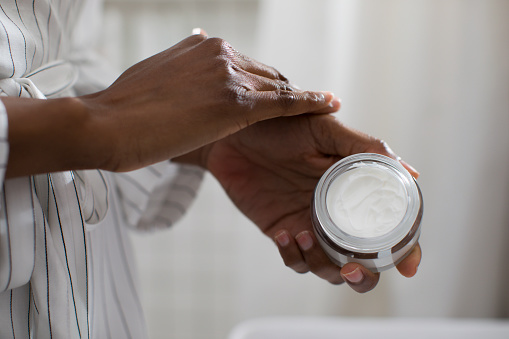 Hands of unrecognisable African woman holding cosmetic cream and applying on her skin.