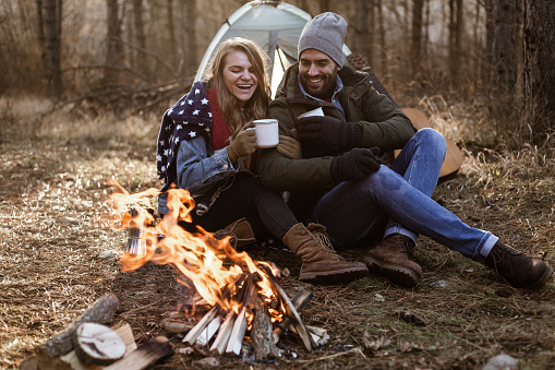 Couple is drinking coffee and sitting in front of a tent next to campfire