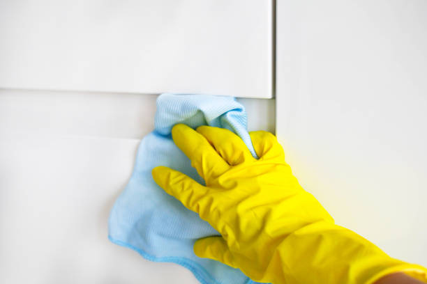 A female hand in yellow rubber gloves wipes a modern white kitchen with a microfiber cloth.