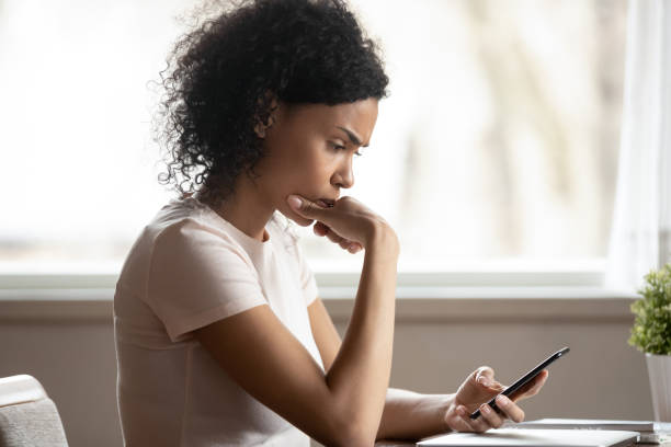 Pensive african American woman lost in thoughts using cellphone Pensive african American millennial female look at smartphone screen thinking of gadget problem, thoughtful biracial woman using modern cellphone reading news, pondering or considering e mail spam photos stock pictures, royalty-free photos & images