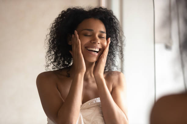 Happy african American woman touch healthy skin in bath Smiling african American woman in towel touch healthy glowing skin look in mirror satisfied with facial treatment procedure, happy biracial young female in bathroom glad with skincare daily routine body care shower stock pictures, royalty-free photos & images