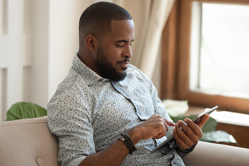 Focused african American millennial guy sit on couch hold tablet browsing internet checking mail, biracial young man rest at home using pad electronic gadget, texting messaging online on device