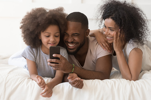 Overjoyed african American family with little daughter have fun in bedroom watch funny video on cellphone together, happy biracial parents enjoy morning cartoons on smartphone with kid laughing