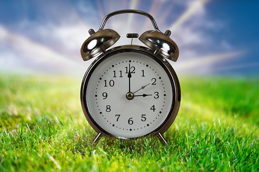 Daylight saving time concept. Alarm clock in green grass with blue sky and sunbeams in background.