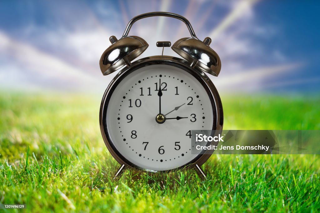 Daylight saving time concept with Alarm clock. Daylight saving time concept. Alarm clock in green grass with blue sky and sunbeams in background. Daylight Saving Time Stock Photo