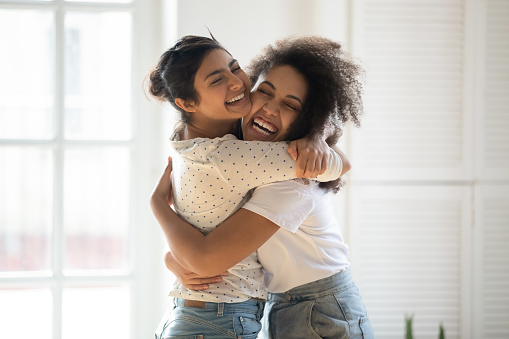 Happy multiethnic millennial girls have fun indoors hugging showing ultimate love and support, smiling multiracial young female friends embrace cuddle feel excited and overjoyed, friendship concept