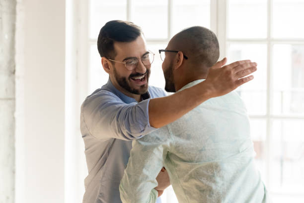 Smiling male friends embrace tap shoulder meeting indoors Smiling Caucasian man in glasses feel overjoyed meeting ethnic male friend or colleague, happy young multiethnic guys hug embrace tapping shoulder excited encounter at party, friendship concept male friendship stock pictures, royalty-free photos & images