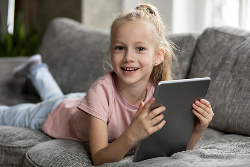 Portrait of smiling little girl lying relaxing on couch in living room using modern tablet at home, happy small child rest on sofa browse Internet play game on pad, children technology concept