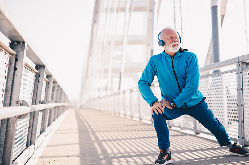 An elderly man stretching outdoors and wearing headphones.