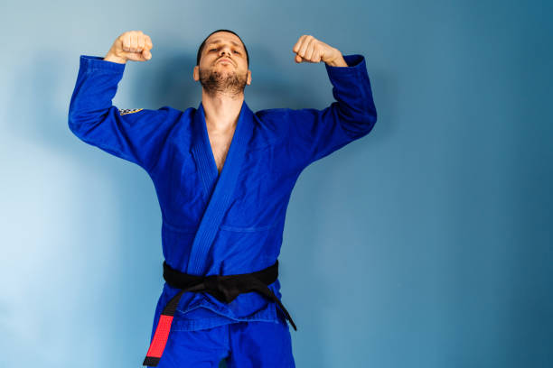 brasilianische jiu jitsu bjj kaukasischen athleten lehrer professor oder kämpfer stehen vor der blauen wand tragen kimono gi einheitliche frontansicht beugen arme zuversichtlich siegreich - ju jitsu stock-fotos und bilder