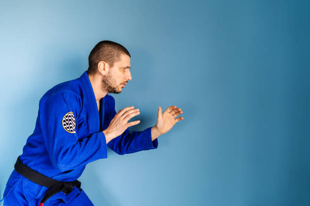 brasilianische jiu jitsu bjj kaukasischen athleten lehrer professor oder kämpfer stehen vor der blauen wand tragen kimono gi einheitliche seitenansicht in kampfhaltung - ju jitsu stock-fotos und bilder