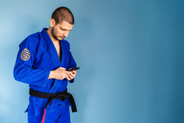 brasilianische jiu jitsu bjj kaukasischen athleten lehrer steht vor der blauen wand tragen kimono gi uniform und schwarzen gürtel aussehende smartphone in den händen halten mit app-anwendung - ju jitsu stock-fotos und bilder