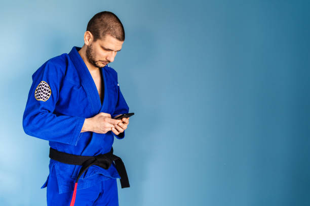 Brazilian jiu jitsu bjj caucasian athlete instructor standing in front of the blue wall wearing kimono gi uniform and black belt looking smart mobile phone in hands holding using app application Brazilian jiu jitsu bjj caucasian athlete instructor standing in front of the blue wall wearing kimono gi uniform and black belt looking smart mobile phone in hands holding using app application brazilian jiu jitsu photos stock pictures, royalty-free photos & images