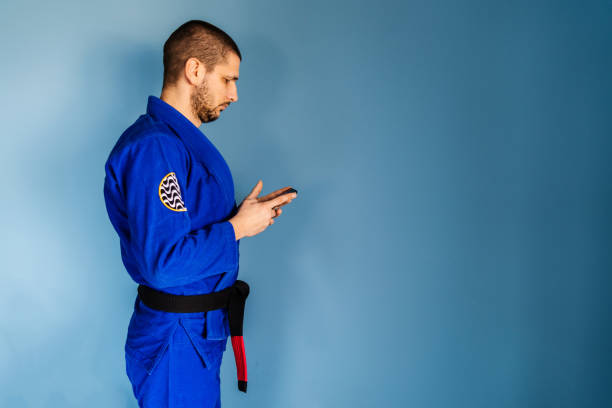 brasilianische jiu jitsu bjj kaukasischen athleten lehrer steht vor der blauen wand tragen kimono gi uniform und schwarzen gürtel aussehende smartphone in den händen halten mit app-anwendung - ju jitsu stock-fotos und bilder