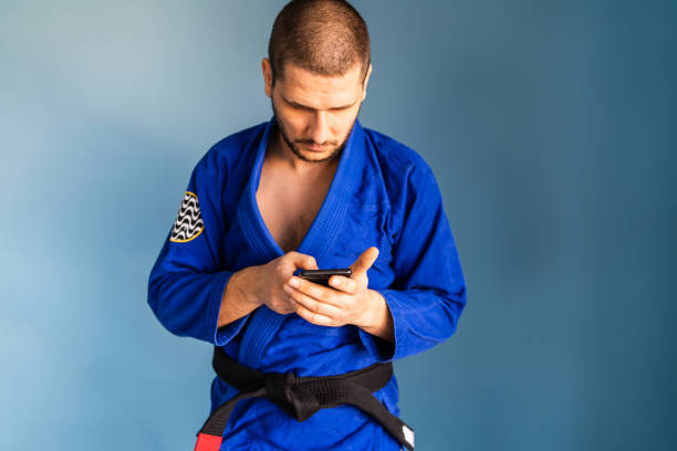 brasilianische jiu jitsu bjj kaukasischen athleten lehrer steht vor der blauen wand tragen kimono gi uniform und schwarzen gürtel aussehende smartphone in den händen halten mit app-anwendung - ju jitsu stock-fotos und bilder