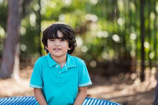 A 6 year old Hispanic boy hanging out at the park. He is sitting on a table, smiling at the camera.
