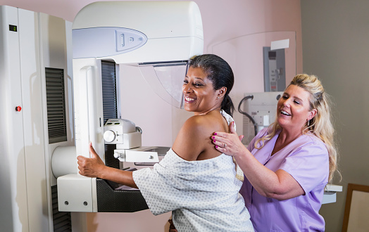 A mature African-American woman in her 40s wearing a hospital gown, getting her annual mammogram.  She is being helped by a technologist, a blond woman wearing scrubs.