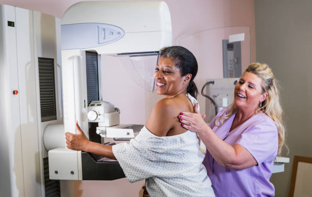 African-American woman getting a mammogram A mature African-American woman in her 40s wearing a hospital gown, getting her annual mammogram.  She is being helped by a technologist, a blond woman wearing scrubs. x ray image medical occupation technician nurse stock pictures, royalty-free photos & images
