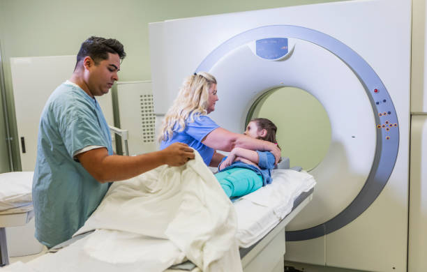 Technologist talking to girl getting CT scan A 7 year old girl getting ready for a CT scan. One of the technologists, a mature woman in her 40s, is talking to the child, explaining the procedure. A mid adult Hispanic man is adjusting the sheets. male nurse male healthcare and medicine technician stock pictures, royalty-free photos & images