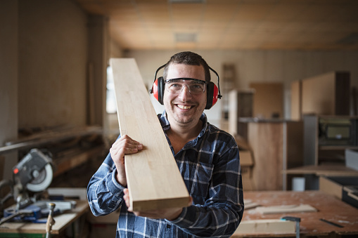 Smiling carpenter holding a wooden beam in workshop, wearing ear muffs and protective goggles, he smiling and looking at camera.