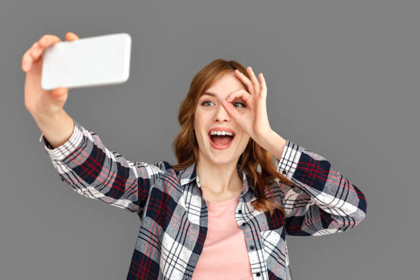 istock Freestyle. Stylish woman studio standing isolated on grey taking selfie on smartphone looking through hand circle smiling cheerful 1201544842