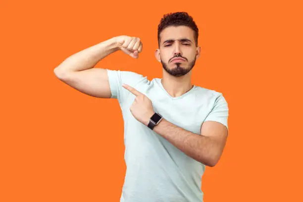 Photo of Look at my strength. Portrait of proud handsome brunette man pointing at biceps. indoor studio shot isolated on orange background