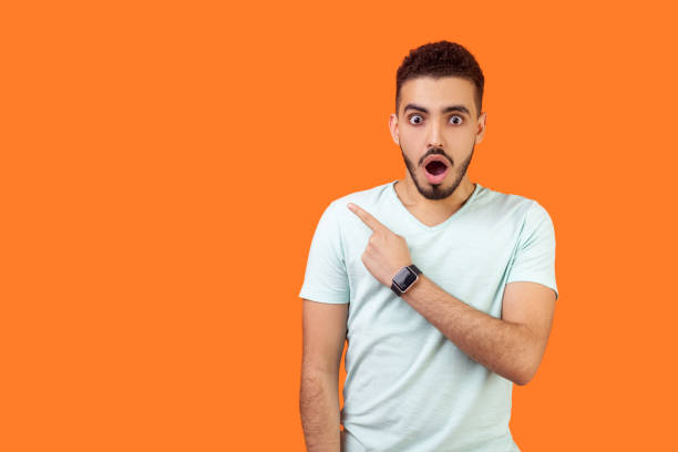 Shocking news! Portrait of astonished brunette man pointing at left. indoor studio shot isolated on orange background Shocking news! Portrait of astonished brunette man with beard in white t-shirt standing with open mouth, looking surprised and pointing at left, empty space for text, advertise. indoor studio shot surprised men stock pictures, royalty-free photos & images