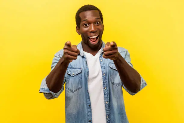 Photo of Hey you! Portrait of happy fascinated man pointing fingers at camera. indoor studio shot isolated on yellow