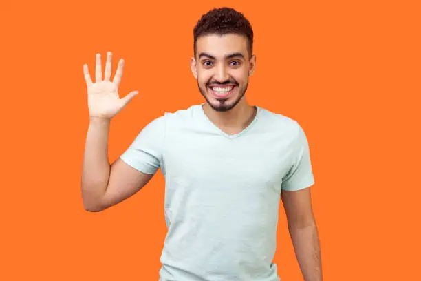 Photo of Hello! Portrait of friendly glad handsome brunette man showing hi gesture. indoor studio shot isolated on orange background