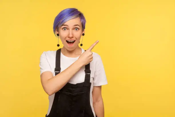 Portrait of astonished surprised fashionable hipster girl with violet short hair in denim overalls pointing to the side freespace for advertising, copy space. isolated on yellow background studio shot