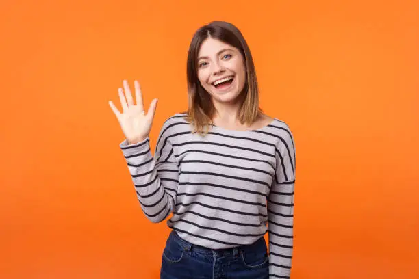 Hello! Portrait of adorable friendly woman with brown hair in long sleeve shirt standing waving hand, looking at camera with engaging toothy smile. indoor studio shot isolated on orange background