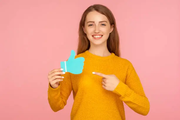 Photo of Like and share! Portrait of attractive happy red hair girl in sweater pointing at blue thumb up love icon and smiling