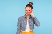 Delighted carefree girl in fashionable outfit peeking through doughnut hole and smiling, having fun with snack, looking into donut