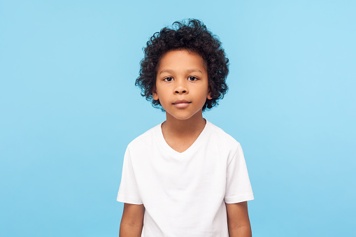 Portrait of cute little boy with stylish curly hairdo in white T-shirt standing, looking at camera with serious attentive face, calm pensive expression. indoor studio shot isolated on blue background