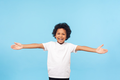 Let's hug. Portrait of friendly hospitable little boy with curls in white T-shirt smiling happily and holding hands wide open to embrace, welcome. indoor studio shot isolated on blue background