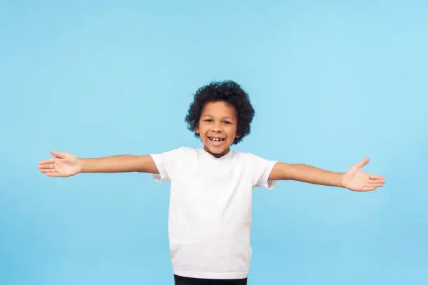 Photo of Let's hug. Portrait of friendly hospitable little boy with curls in white T-shirt smiling happily and holding hands wide open to embrace