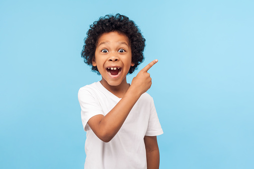 Wow look, advertise here! Portrait of amazed cute little boy with curly hair pointing to empty place on background, surprised preschooler showing copy space for promotional ad. indoor studio shot