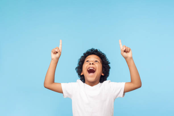 ¡guau, mira hacia arriba! retrato de niño asombrado señalando hacia el lugar vacío sobre fondo azul, expresando el choque - marketing commercial sign african descent african ethnicity fotografías e imágenes de stock