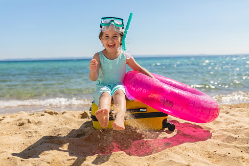 Beautiful smiling child at a beach with a inflatable ring, a suitcase and a skuba mask.