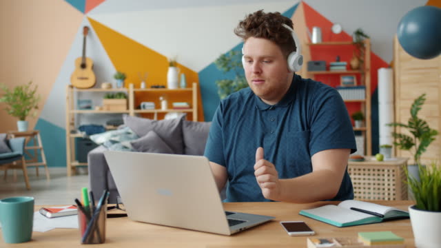 Cheerful guy in headphones listening to music dancing working with laptop at home