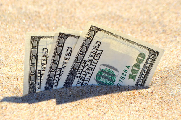istock Money dolars half covered with sand lie on beach close-up. 1201597576