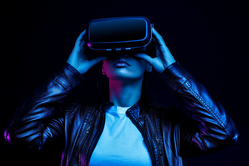Chica afroamericana en gafas vr, viendo vídeo de 360 grados con auriculares de realidad virtual aislados sobre fondo negro, iluminados por luces de neón photo