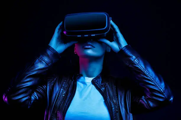 Photo of African american girl in vr glasses, watching 360 degree video with virtual reality headset isolated on black background, illuminated by neon lights