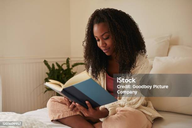 Young woman sitting on her bed reading a novel